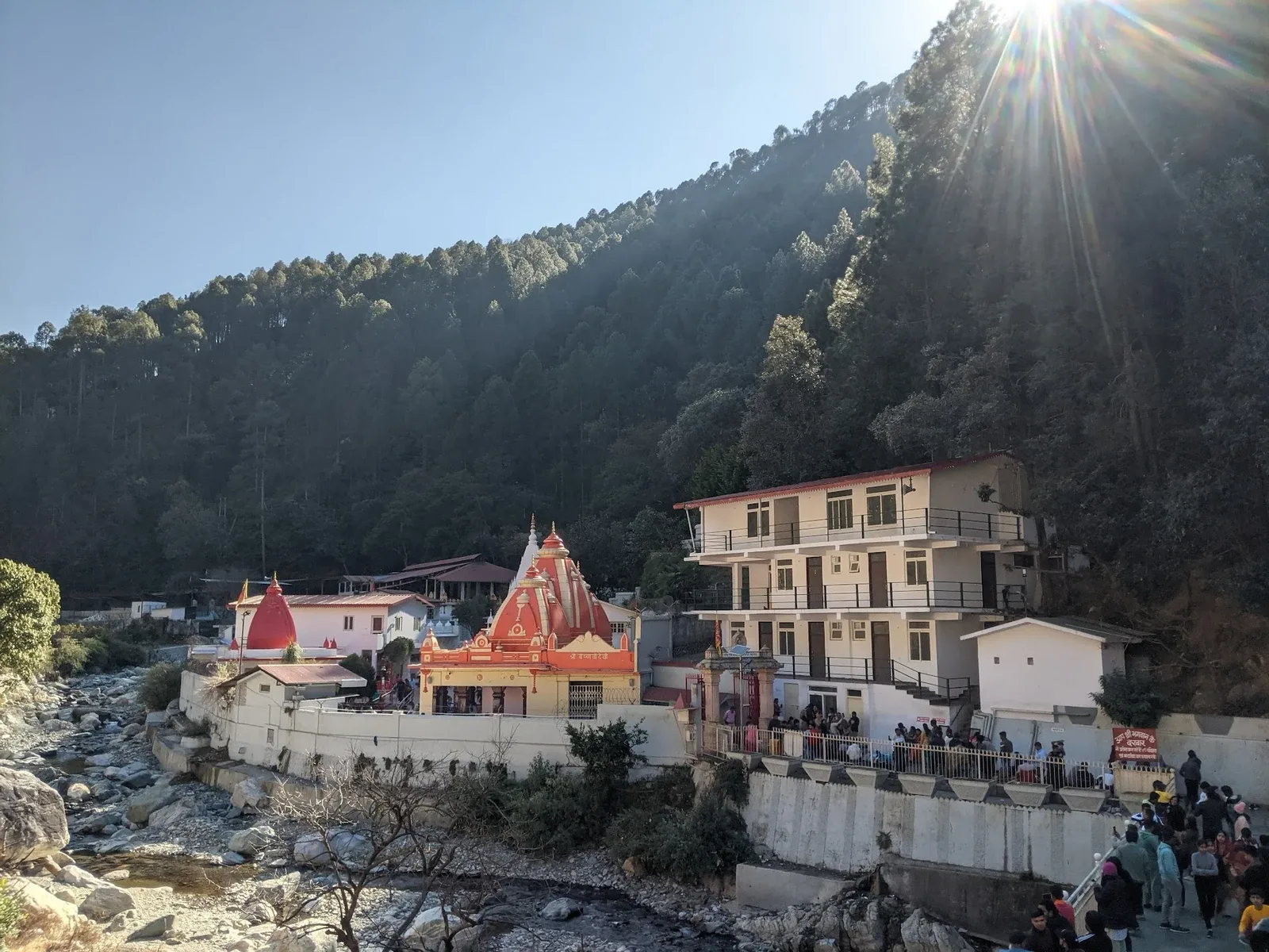 Kainchi Dham temple buildings beside the Kshipra river and a wooded hillside in Nainital district, Uttarakhand.